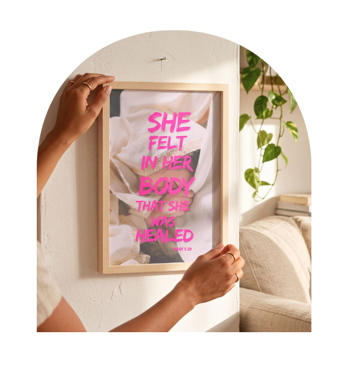 A framed inspirational wall art print with a pink quote over a white and gray abstract background, being held by two hands against a textured off-white wall with a plant and sofa in the background.
