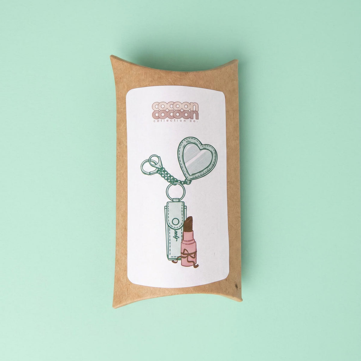 A small kraft paper gift box with a white label printed with an illustration of a compact mirror, keychain, and lip balm, set against a solid mint green background.