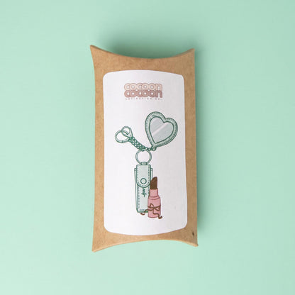 A small kraft paper gift box with a white label printed with an illustration of a compact mirror, keychain, and lip balm, set against a solid mint green background.