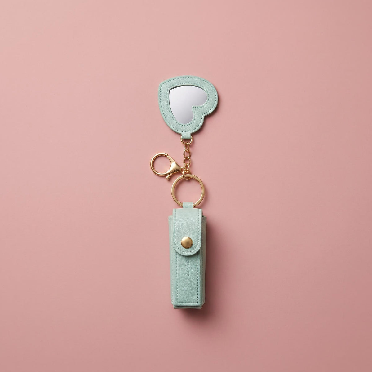 A mint green faux leather keychain organizer with a small heart-shaped mirror and gold-tone hardware, displayed on a solid pink background.