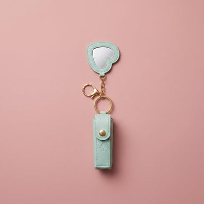 A mint green faux leather keychain organizer with a small heart-shaped mirror and gold-tone hardware, displayed on a solid pink background.