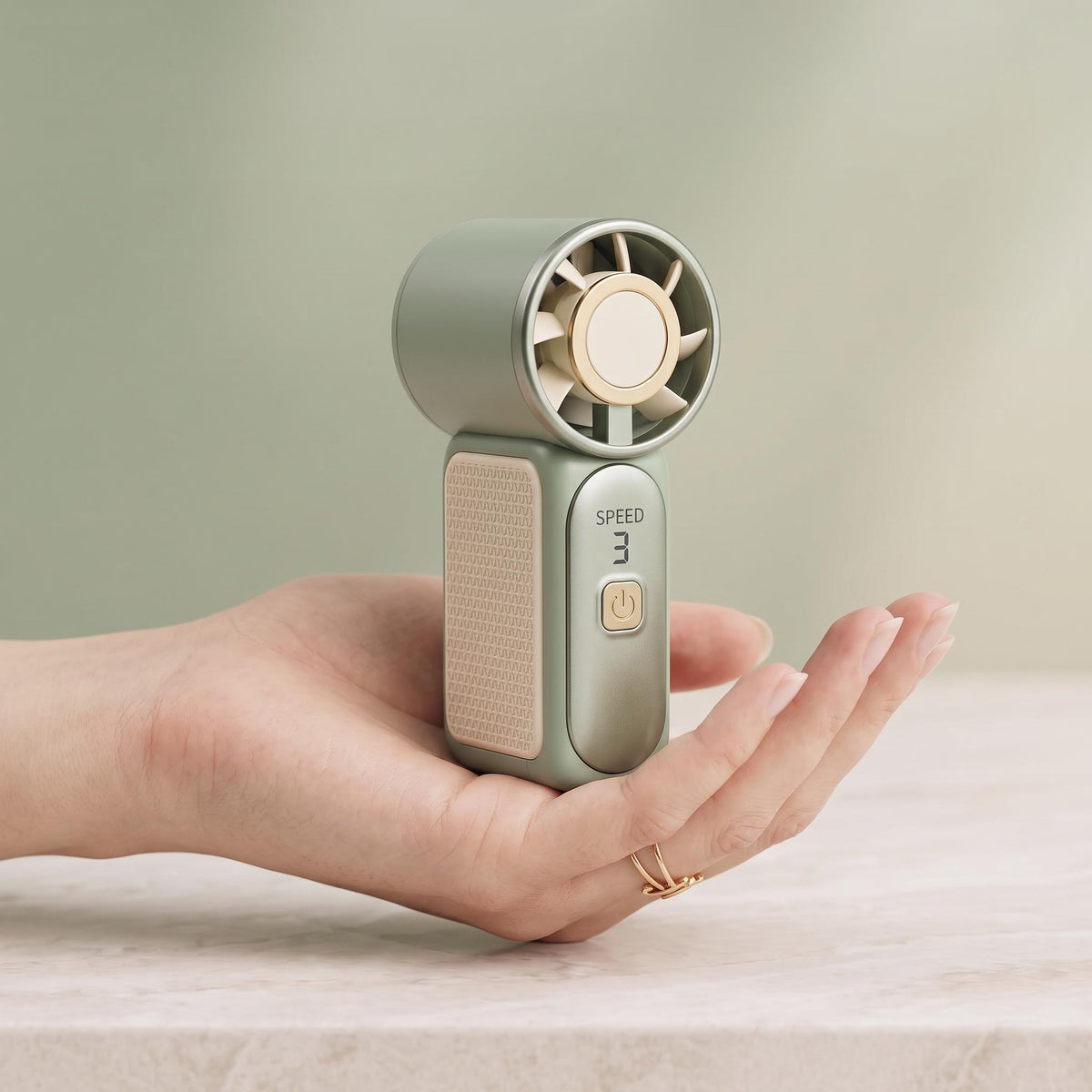 A handheld mini fan in a muted sage green and beige color, being held in a person's hand against a soft, neutral background.