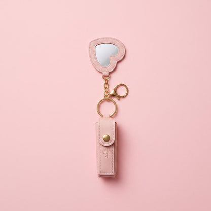 A pink faux-leather keychain organizer with an attached heart-shaped mirror and gold-tone hardware, displayed on a solid pink background.