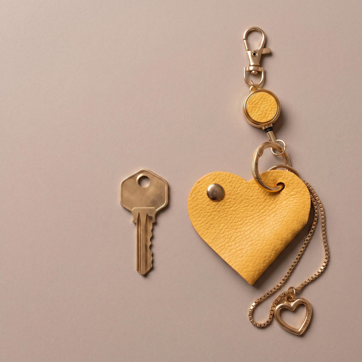A yellow textured heart-shaped keychain with a gold-tone clasp, extension ring, chain, and small heart pendant, shown next to a metal key on a plain warm gray background.