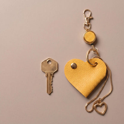A yellow textured heart-shaped keychain with a gold-tone clasp, extension ring, chain, and small heart pendant, shown next to a metal key on a plain warm gray background.