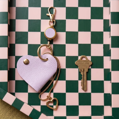 A lavender faux-leather heart-shaped keychain with gold-tone hardware, shown next to a metal key on a green and pink checkered tray.