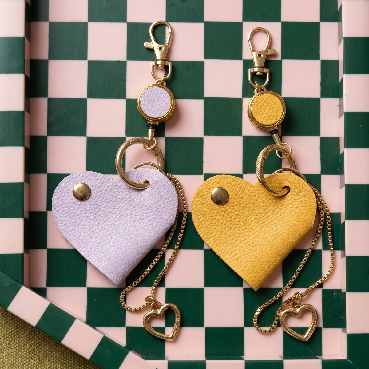 Two heart-shaped leather keychains, one light lavender purple and one mustard yellow, with gold-tone hardware, displayed on a green and pink checkered tray.