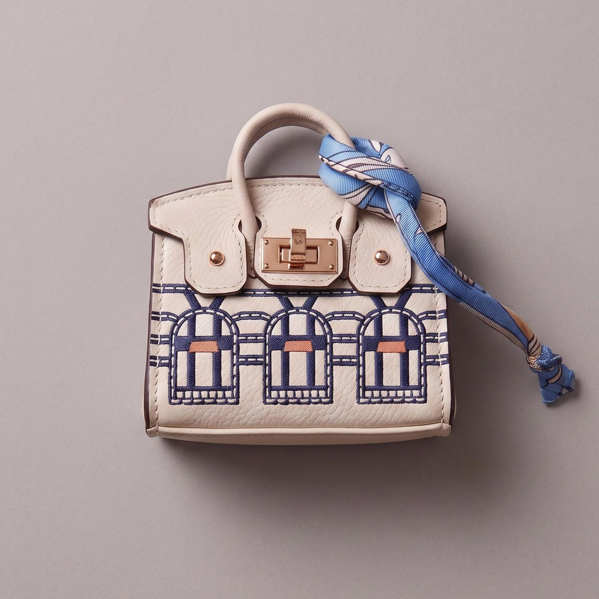 A small structured handbag in off-white leather with a blue and navy architectural pattern on the lower front, a gold-tone turn-lock closure, and a blue patterned scarf tied around the handle, photographed on a plain light gray background.