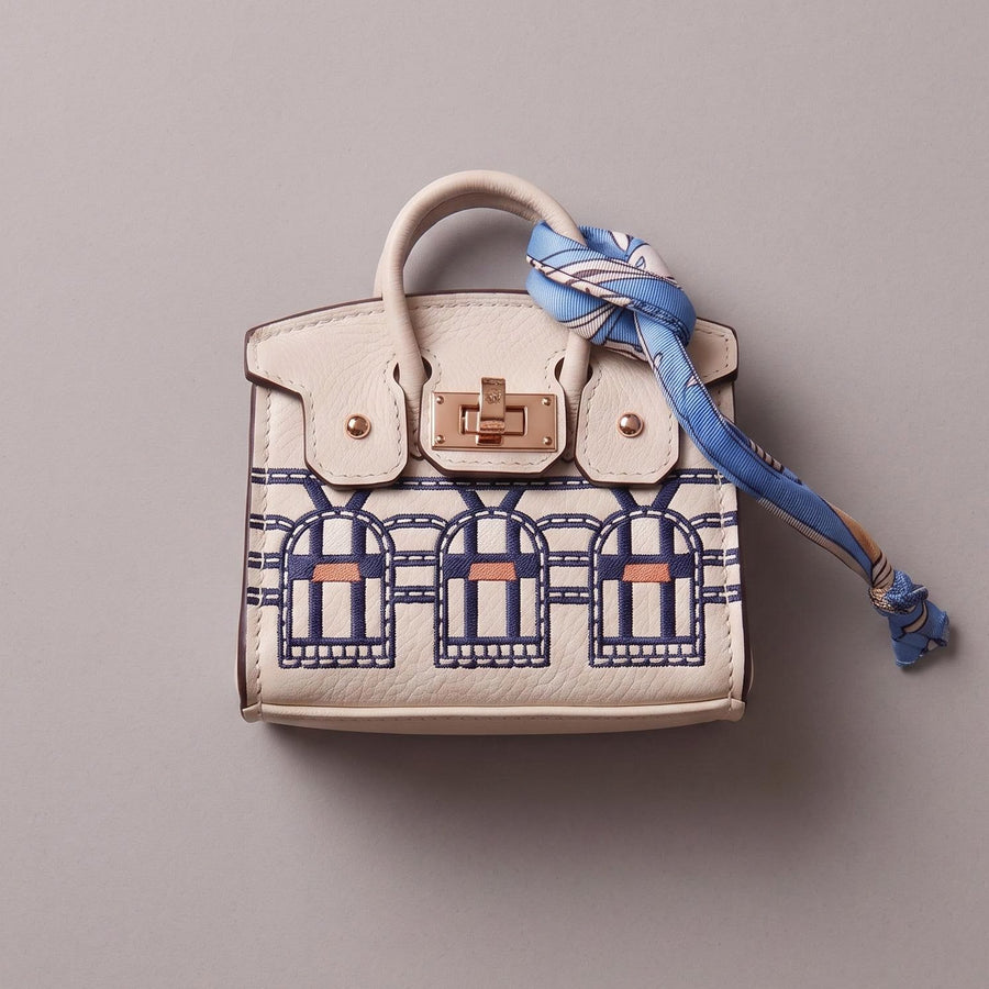 A small structured handbag in off-white leather with a blue and navy architectural pattern on the lower front, a gold-tone turn-lock closure, and a blue patterned scarf tied around the handle, photographed on a plain light gray background.